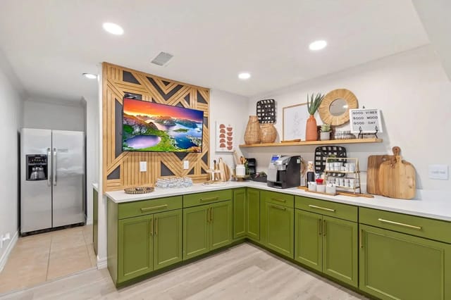 Modern communal kitchenette with green cabinets, a coffee station, and TV on a geometric wood panel.