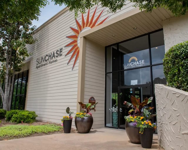 Exterior entrance of Sunchase Apartment Homes with glass doors and sunburst logo, surrounded by potted plants.