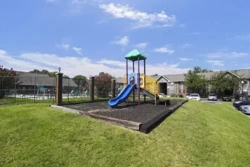 Playground with a blue slide and climbing structure in a grassy, fenced apartment complex courtyard.