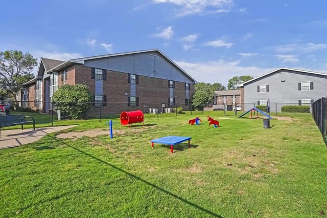 Grass-covered community playground with fencing and playground equipment beside apartment buildings.