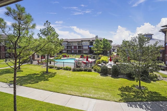 Outdoor apartment community pool area with lounge chairs and red umbrellas, surrounded by trees.