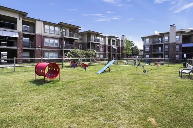 Courtyard of an apartment complex with a fenced playground, benches, and green lawn.