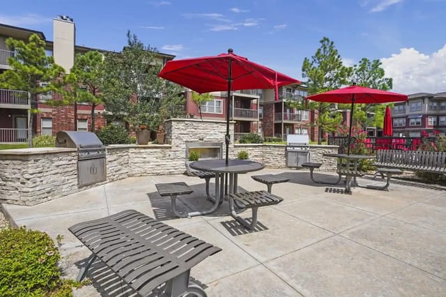 Outdoor communal courtyard with grills, tables, benches, and red umbrellas near apartments.