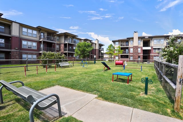 Apartment complex with a fenced-in dog park area in the foreground, featuring agility equipment and benches.