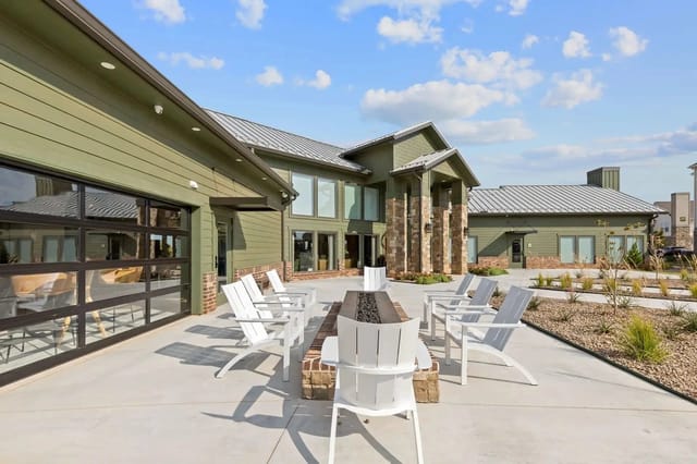 Patio area with white chairs around a fire pit in front of a green building.