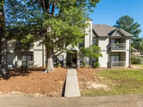 Exterior view of a two-story apartment building with balconies and a central walkway among trees.