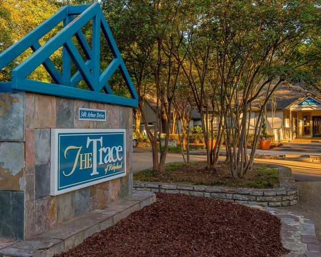 Exterior entrance sign reading The Trace with landscaping and a building in the background.