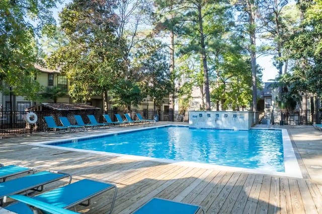 Outdoor apartment community pool with blue lounge chairs and surrounding trees.
