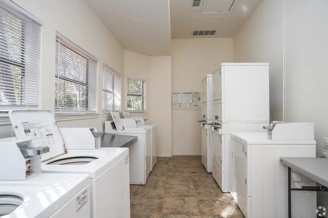 Row of white washing machines and dryers in a bright communal laundry room.