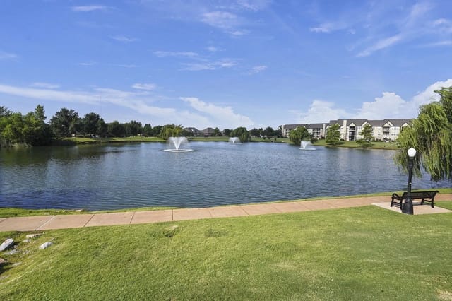Pond with fountains, grassy park, and a bench along a walkway with apartment buildings in the distance.