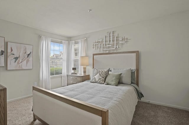 Neutral-toned bedroom in an apartment with a bed, nightstand, and large window with white curtains.