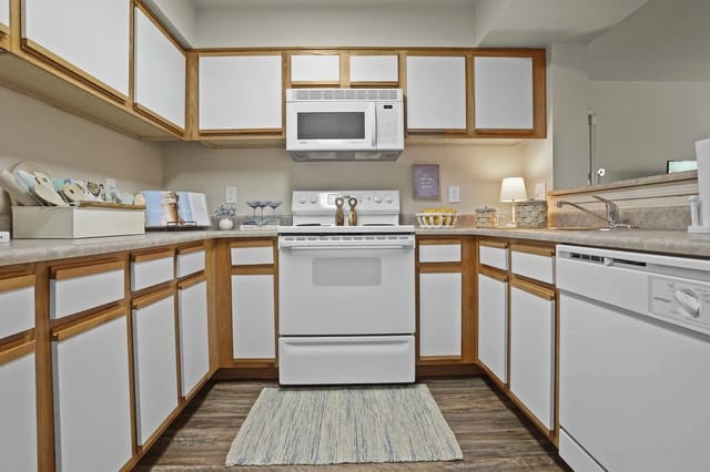 U-shaped apartment kitchen with white cabinets, wood trim, and white appliances.