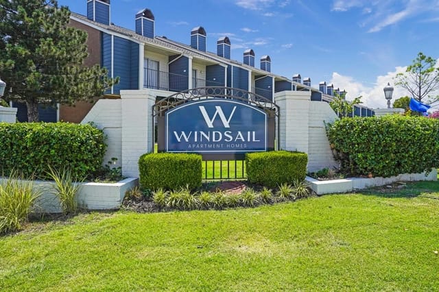Entrance sign for Windsail Apartment Homes with shrubs and a blue building behind.