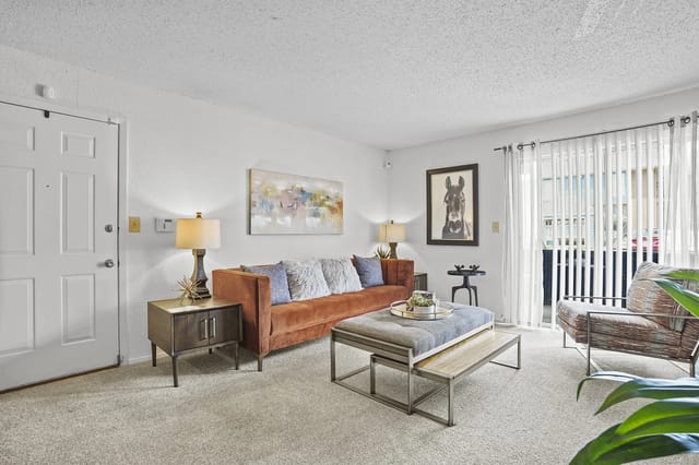 Bright living room in an apartment with a tan sofa, coffee table, art, and large window with vertical blinds.