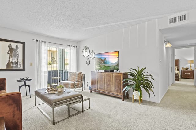 Apartment living room with TV on a wooden stand, seating, plant, and sliding door to balcony.