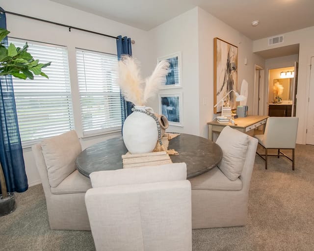 Living/dining area in an apartment with a round glass table and beige upholstered chairs.