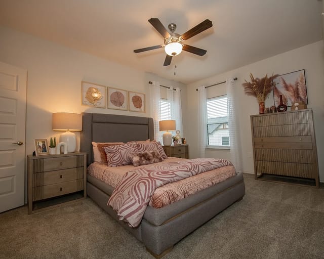 Bedroom with gray upholstered bed, two nightstands, dresser, ceiling fan, and two windows.
