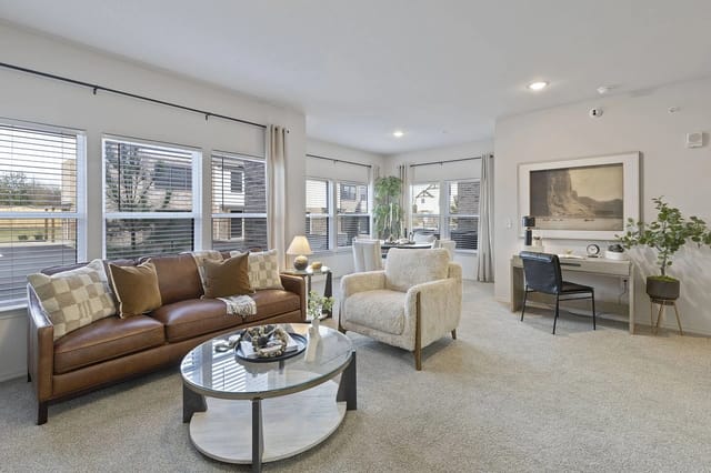 Bright living room with large windows, a brown leather sofa, cream chair, and glass coffee table.