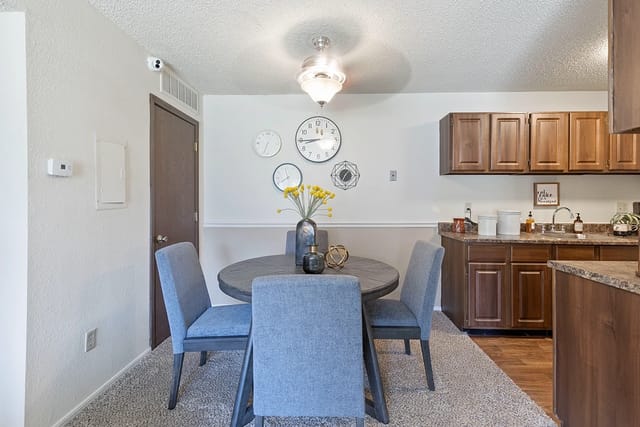 Dining area in apartment with round table, four blue chairs, and a kitchen with brown cabinets.