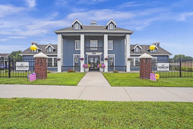 Front entrance of a blue apartment community building with a gate, pillars, and manicured lawn.