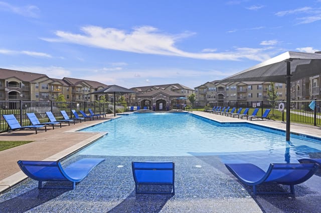 Resort-style swimming pool with blue lounge chairs and apartment buildings in the background.