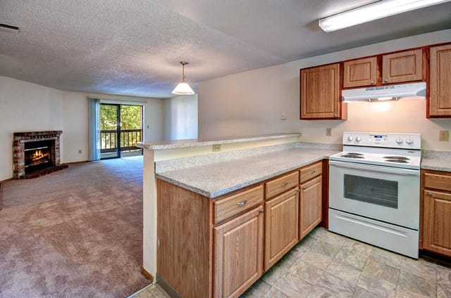 Kitchen with wood cabinets, white stove, and a pass-through counter to the living room with a fireplace.