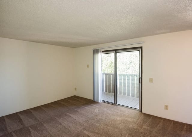 Empty living room with sliding glass door to balcony, beige carpet, white walls.