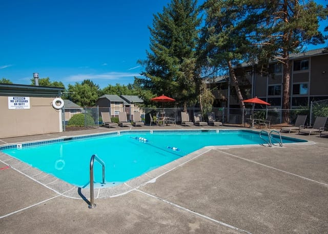 Outdoor apartment pool with lounge chairs, red umbrellas, and a surrounding fence.