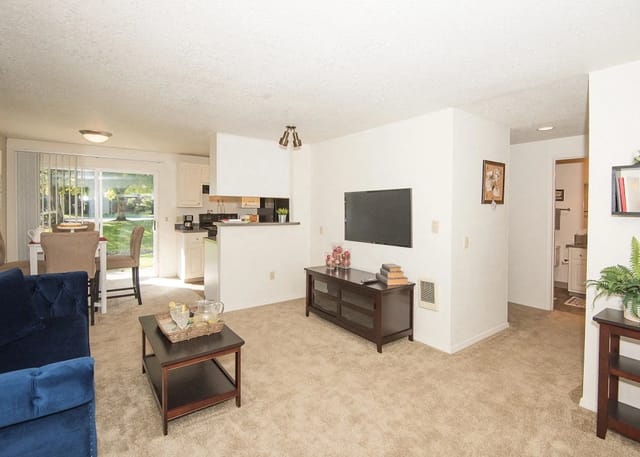 Living room with beige carpet, white walls, and a TV console, open to a small kitchen and dining area.