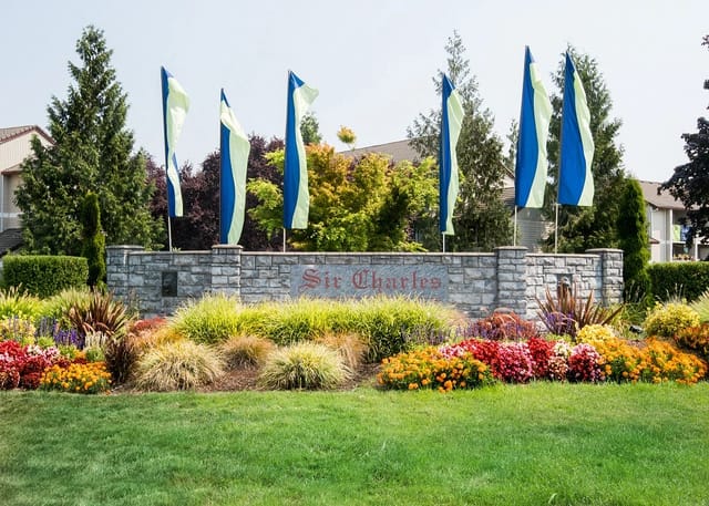 Stone entry sign reading 'Sir Charles' with blue-green flags and colorful flowerbeds.