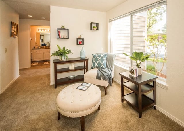 Cozy apartment living room with beige carpet, ottoman, chair, shelves, and a large window.