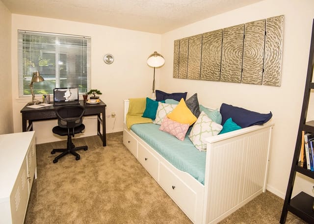 Small apartment bedroom with a white daybed, dark desk by the window, swivel chair, and a tall bookshelf.
