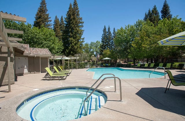 Outdoor pool and circular hot tub with green lounge chairs and trees.