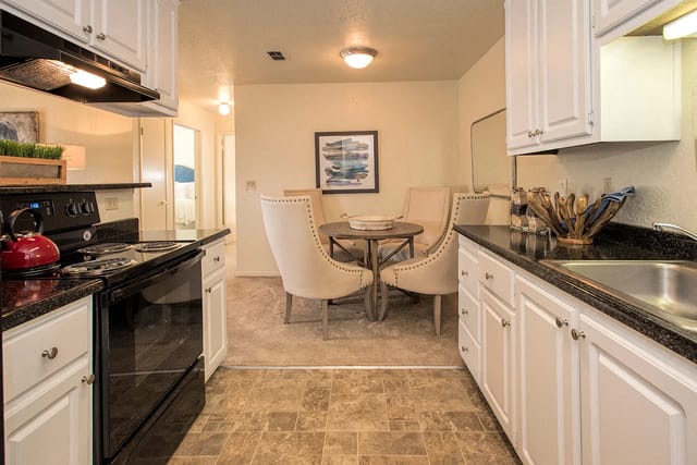 Open kitchen and dining area in an apartment, with white cabinets, black countertops, and a round table with beige upholstered chairs.