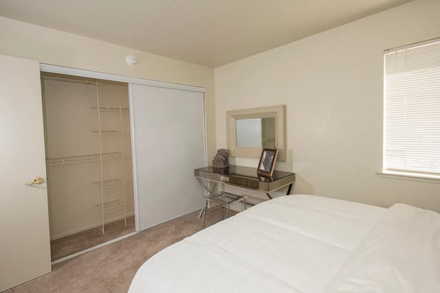 Bedroom in an apartment with a sliding closet door, dresser, mirror, and window blinds.