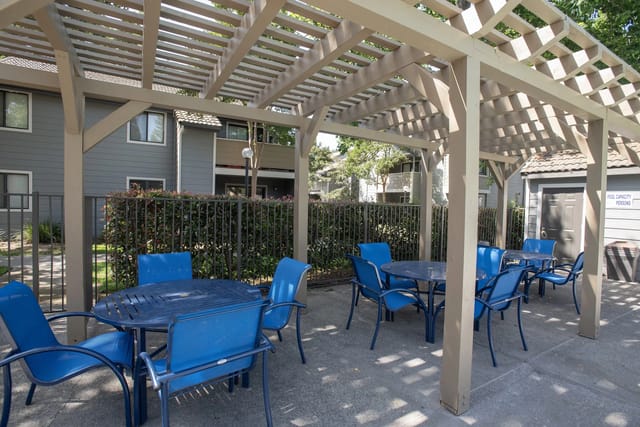 Outdoor communal seating area with blue chairs and tables under a wooden pergola.