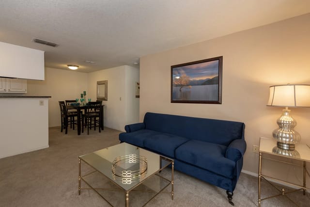 Living room in an apartment with a blue sofa, glass coffee table, lamp, and a dining area.