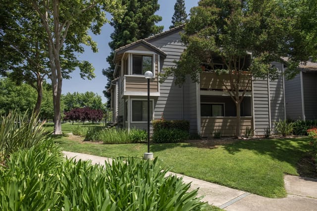 Exterior view of a gray apartment building with landscaped lawn, trees, and a sidewalk.