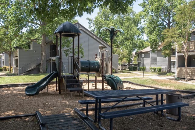 Playground with slides and climbing structure in a tree-lined apartment community courtyard.