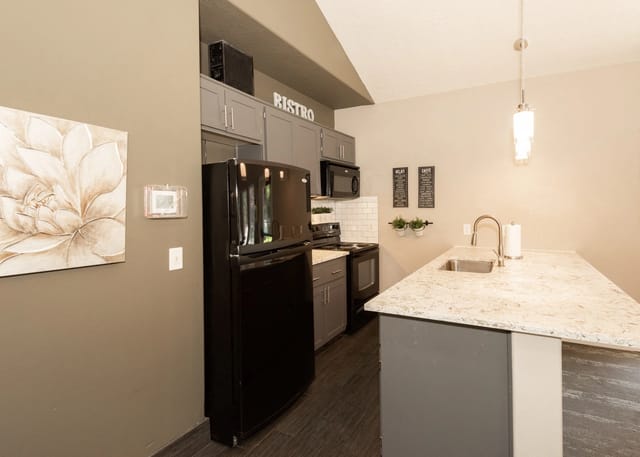 Modern kitchen with a large black refrigerator, grey cabinets, and a granite island with a sink.