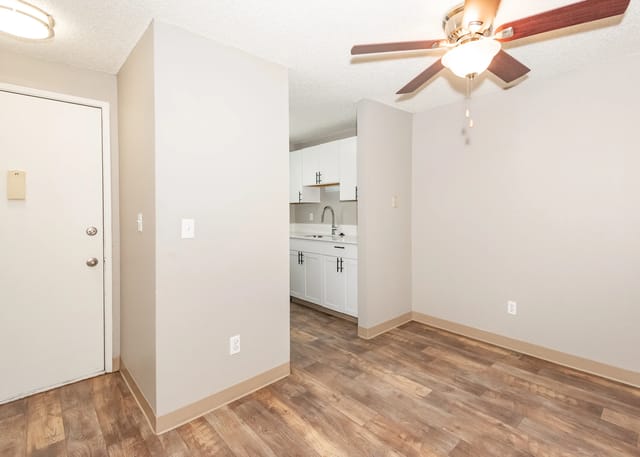 Interior of an apartment with an entryway opening to a small kitchen with white cabinets and a ceiling fan.