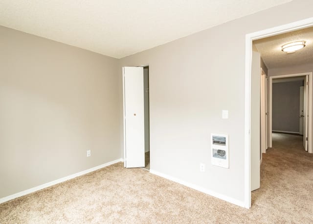 Empty beige bedroom with carpet, a partially open closet door, and a hallway visible.