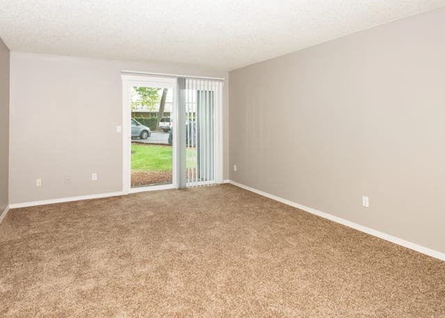 Empty living room with beige walls, brown carpet, and a sliding glass door to outdoors.