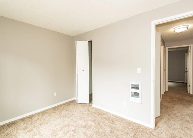 Empty room with beige carpet and light gray walls, view into a hallway.