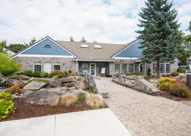 Stone building with blue siding and large windows, surrounded by landscaping and a decorative bridge.