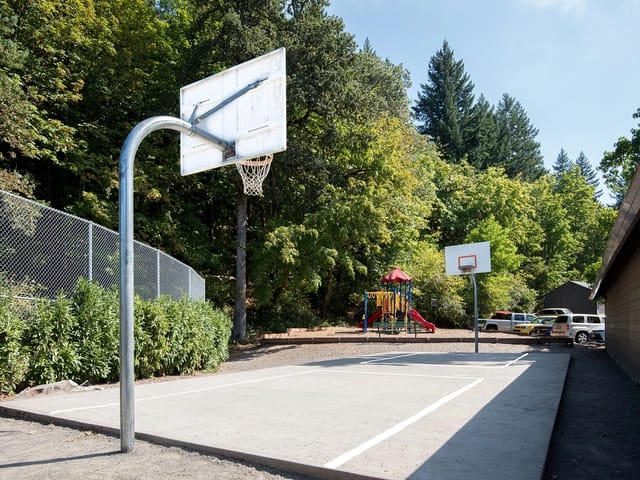Outdoor basketball court with two hoops, a playground, and trees in the background.