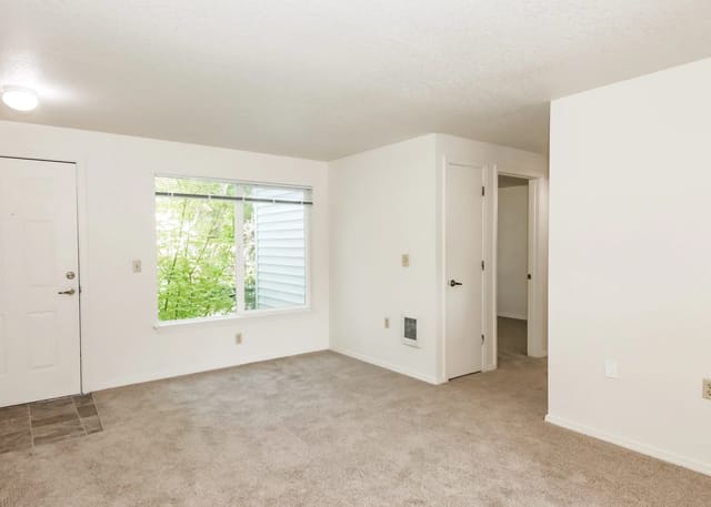 Empty living room with carpet, a window overlooking trees, and two doors.