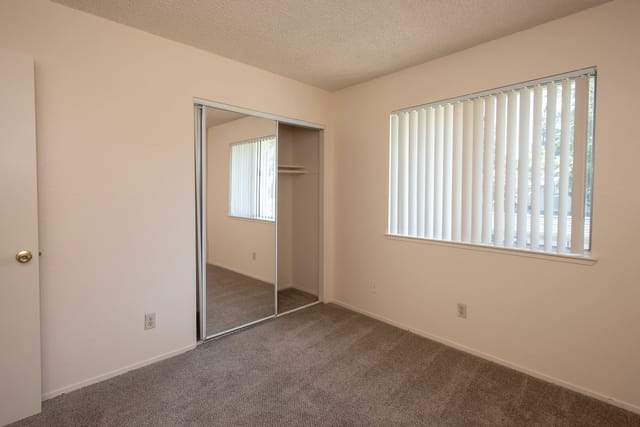 Bedroom with mirrored closet doors and a large window with vertical blinds.