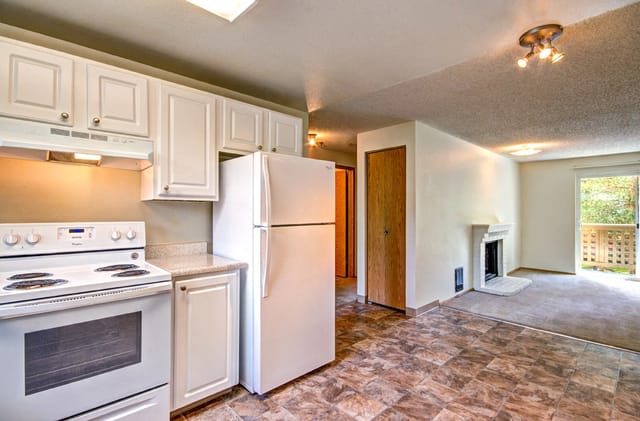 Kitchen with white cabinets, refrigerator, and oven, opening into a living area with a fireplace and balcony access.
