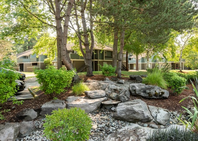 Apartment building exterior with mature trees and a rocky landscape in the foreground.